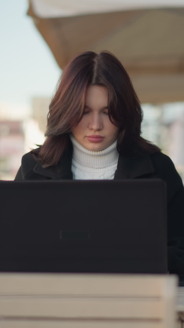 dama trabajando en una computadora portátil al aire libre en un entorno urbano, con un elegante abrigo negro, con un pilar negro decorativo cerca, un edificio de ladrillo rojo borroso en el fondo, y un coche pasando en el borrón