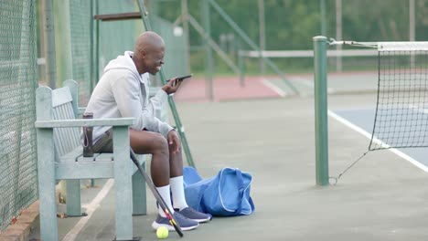 happy african american man talking on smartphone sitting on bench at tennis court, slow motion