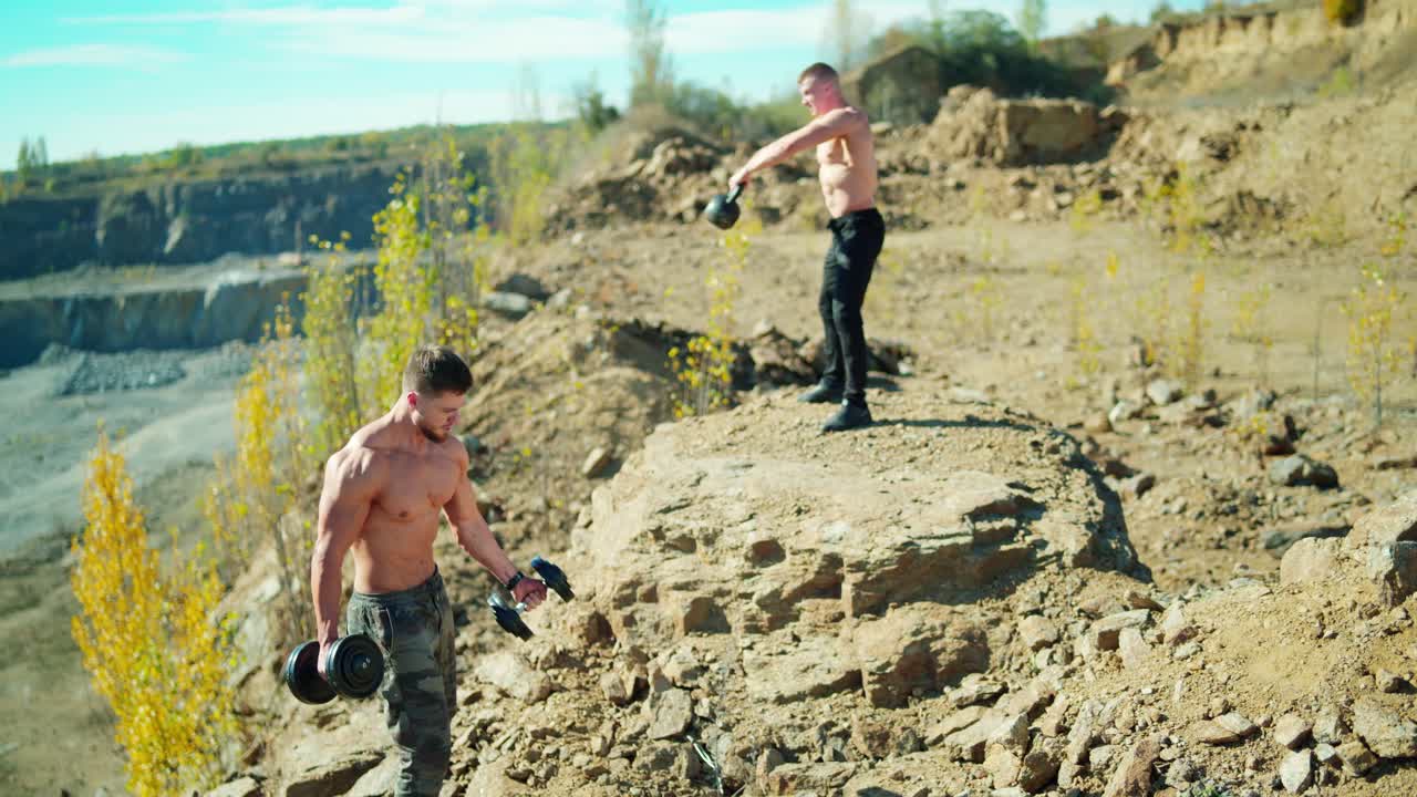 Two athletes training with sports equipment. Muscular man lifting dumbbells outdoors. Strong sportsman lifting kettlebell on the rocky hill.