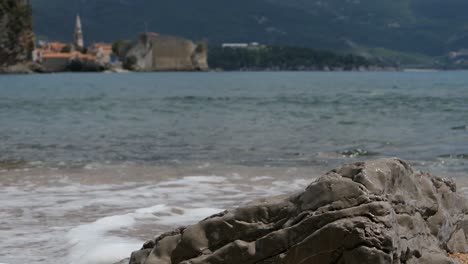 Waves-crashing-on-beach,-closeup,-Budva-Old-Town-background,-Montenegro