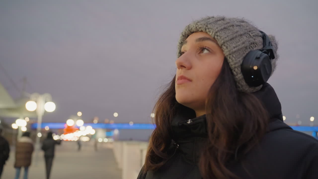 Passeggiata serale con umore riflessivo. Donna contemplativa che si gode il crepuscolo contro il porto e figure distanti. Donna con le cuffie osserva pensierosa le luci del porto durante una tranquilla passeggiata serale.