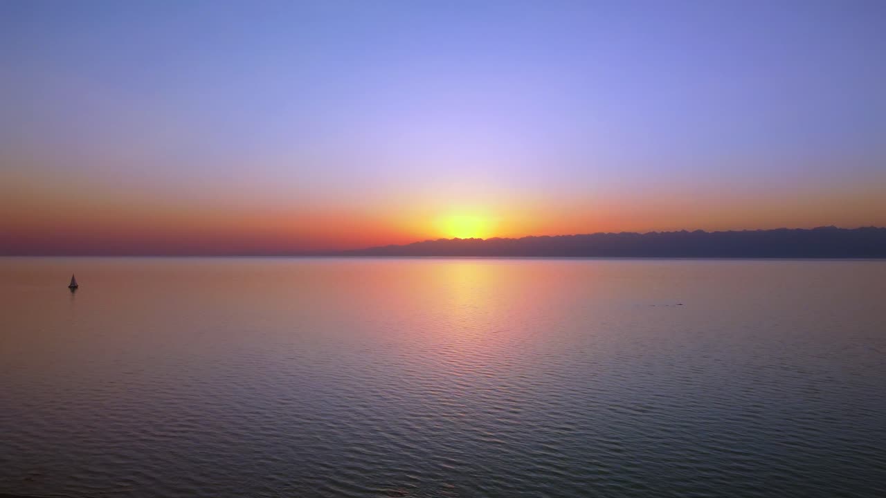 imágenes aéreas de un gran lago capturado durante la puesta de sol, los tonos vibrantes del sol poniente, imponentes montañas en el fondo y un velero activo en el centro