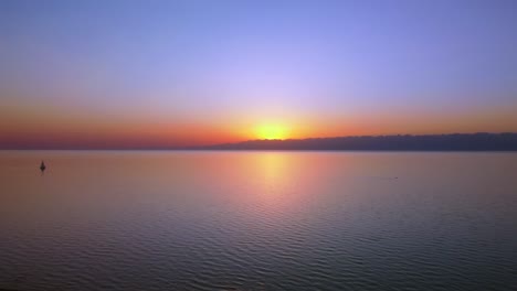 aerial footage of a large lake captured during sunset, the vibrant hues of the setting sun, towering mountains in the background and an active sailing boat in the centre