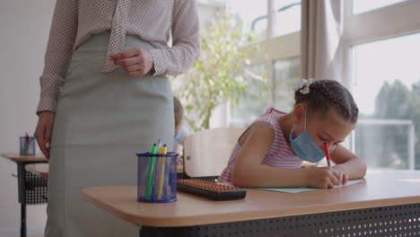 multiracial group of kids wearing face masks working at class writing and listening explanations of teacher in classroom