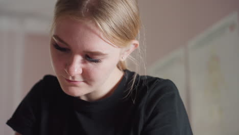 close-up of light skin woman with blonde hair wearing black shirt and smiling subtly while looking downward, surrounded by soft natural light and pastel blurred background in relaxed indoor setting