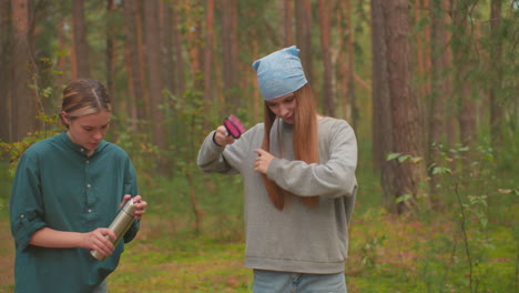 young women pause in forest, one holding thermos and other brushing her hair with a brush, their backpacks rest on ground, and they appear relaxed, enjoying moment of calm in natural setting