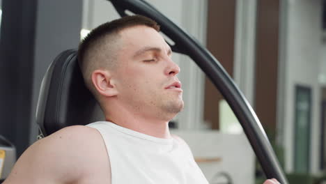 portrait view of man seated on weight bench gripping chest press handles tightly while moving them with strength, eyes closed and face muscles tensed, showcasing focused workout effort in modern gym