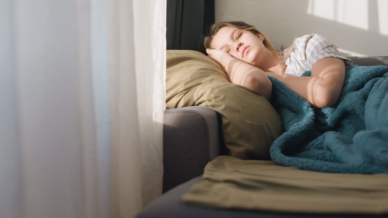 Child Relaxing On Sunlit Sofa, Youthful Student Reclining Comfortably With Cozy Blanket And Pillow, Young Person Takes Restful Break In Serene Sunlit Living Space With Cushion And Warmth