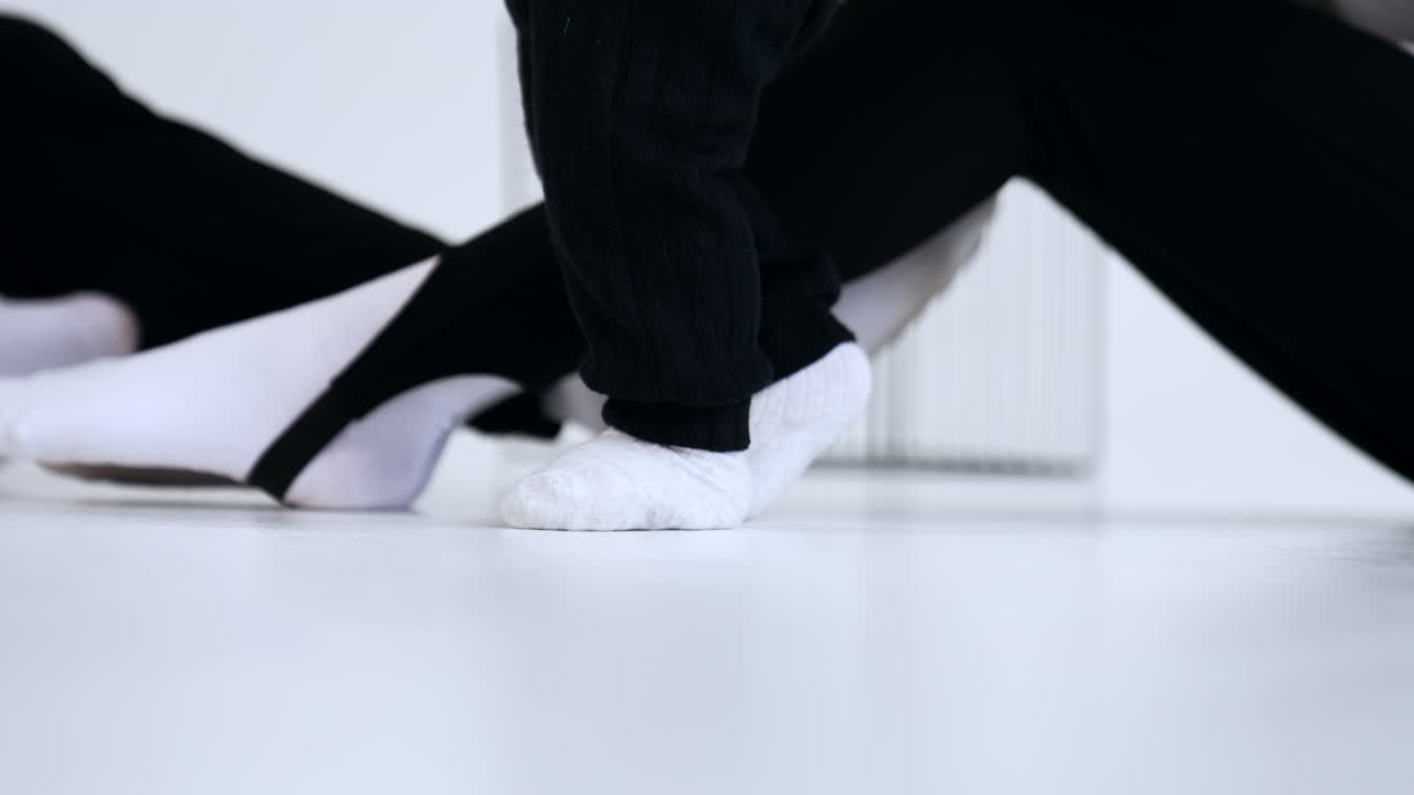 Little feet of a baby in white socks and black pants step by the floor. Legs of parents are at backdrop.