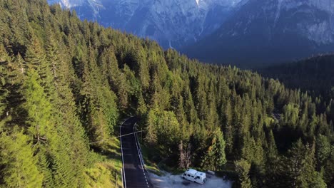 Eine-Drohnenaufnahme-Einer-Bezaubernden-Bergstraße-In-Den-Dolomiten,-Mit-Einem-Autocamper,-Der-Am-Straßenrand-Inmitten-Eines-Von-Bergen-Umgebenen-Pinienwaldes-In-Norditalien-Geparkt-Ist