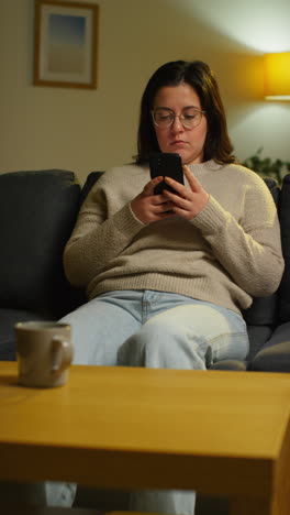 vertical video of young woman spending evening at home sitting on sofa with mobile phone scrolling through internet or social media 1