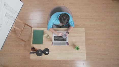 top view of a man using smartphone while sitting at the wooden desk besides a whiteboard typing on a laptop at home