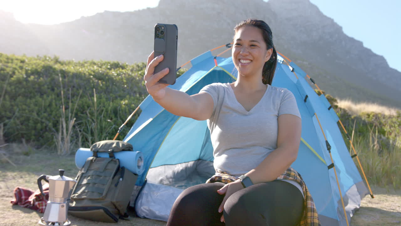 Taking selfie, woman sitting in front of tent during mountain hike