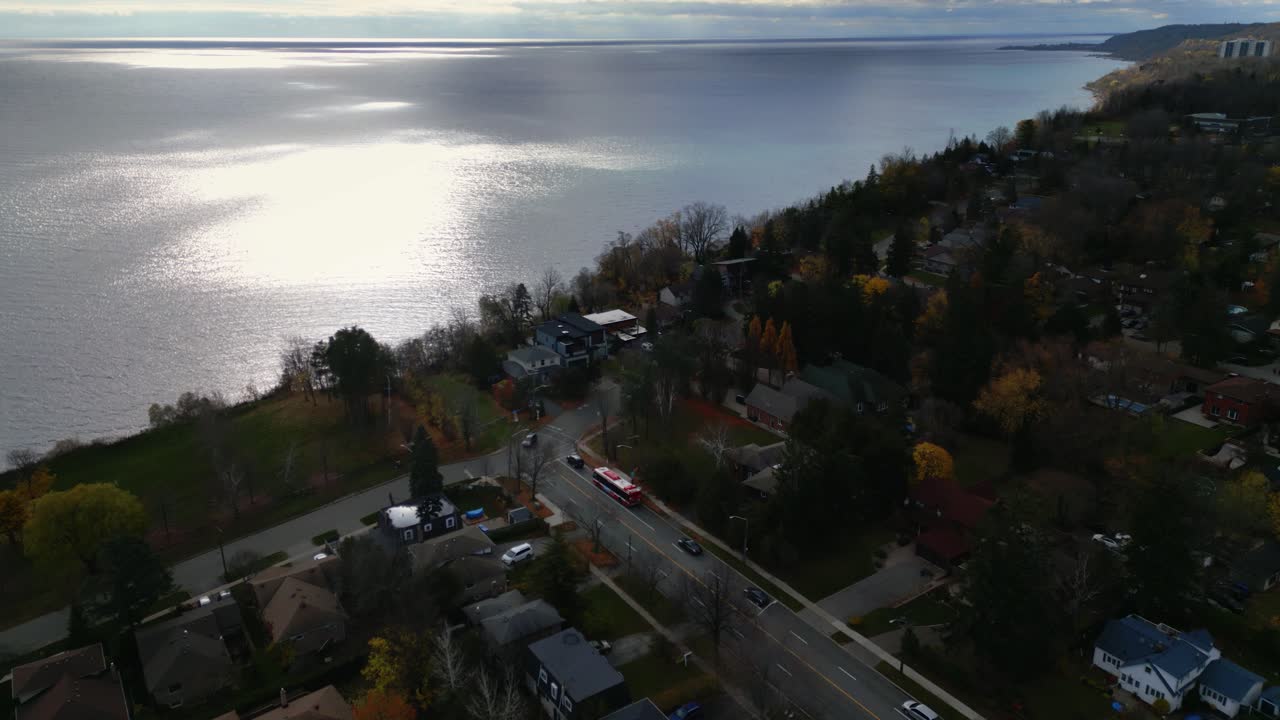 Aerial dolly through suburban area surrounded by hills and trees under cloudy skies, peaceful calm ocean waters as cars enter town