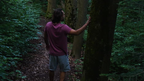 young man in red tshirt and long brown hair walking barefoot down a small path in a green drakish forest and touches some trees