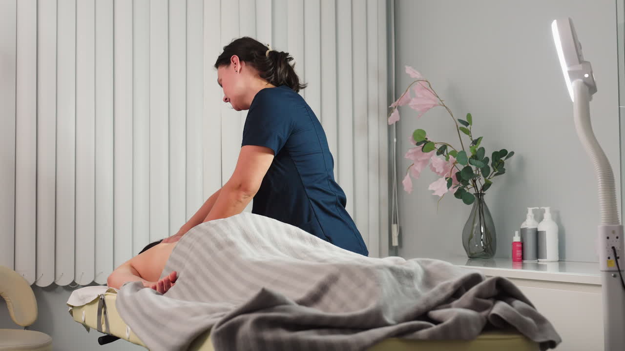 Facialist with hair packaged massaging back of guest on massage bed in spa room with flower vase and massage cream on shelf creating relaxing self care moment framed by soft lighting