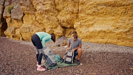 A-brunette-man-in-a-gray-T-shirt-and-a-girl-with-a-bob-hairstyle-in-a-blue-jacket-are-taking-apart-their-backpacks-and-preparing-to-climb-the-yellow-rocks,-they-are-taking-out-the-necessary-ammunition-and-preparing-for-rock-climbing