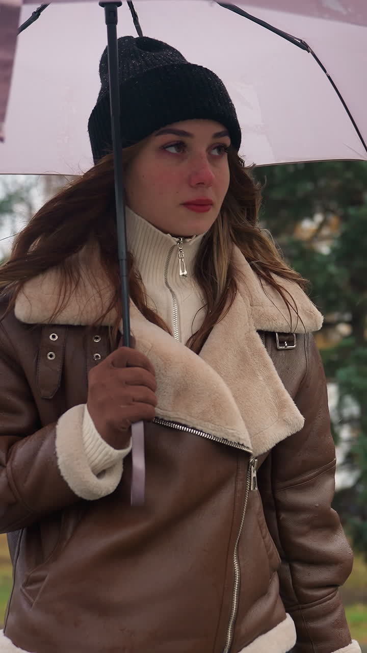 Young woman holding umbrella while walking in park on cold overcast day, wearing black knit cap and brown shearling jacket, surrounded by green pine trees and soft urban landscape with light snowfall