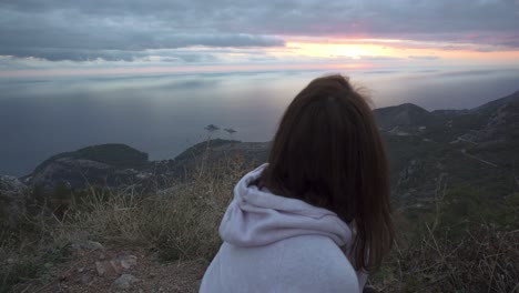 chica en cuclillas en un acantilado alto y ver la puesta de sol a través del cielo nublado sobre el mar adriático