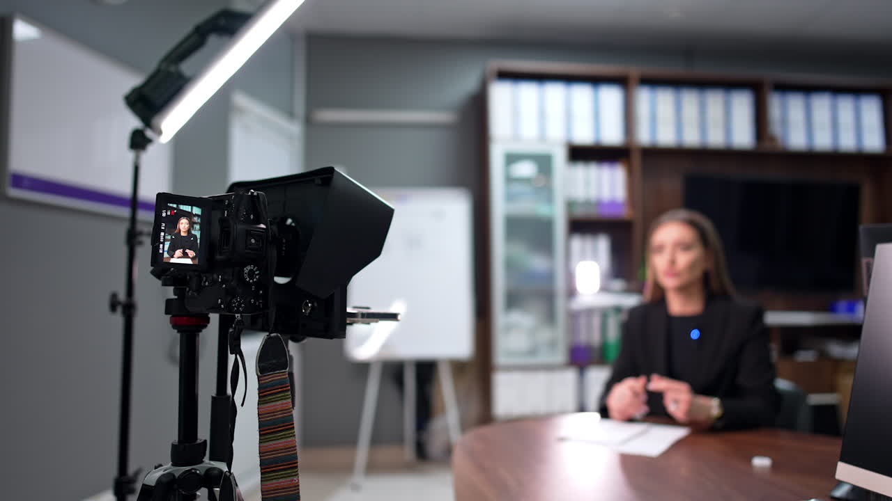 Female brunette talks on the display of professional camera. Positive lady in black jacket takes video for her internet blog. Blurred backdrop.