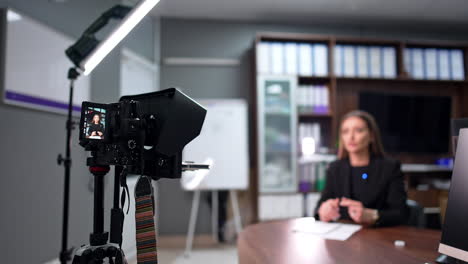 female brunette talks on the display of professional camera. positive lady in black jacket takes video for her internet blog. blurred backdrop.
