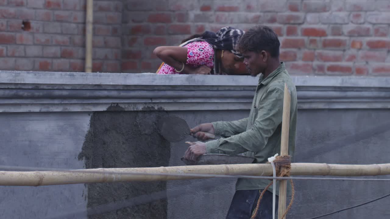 A slow motion handheld long wide shot of an Indian woman wearing pink Kurta and scarf covered on head and a man wearing faded olive green shirt, working at a construction site, applying cement on wall