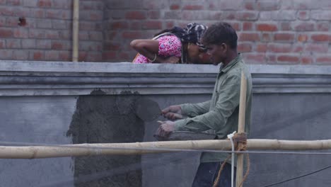 a slow motion handheld long wide shot of an indian woman wearing pink kurta and scarf covered on head and a man wearing faded olive green shirt, working at a construction site, applying cement on wall