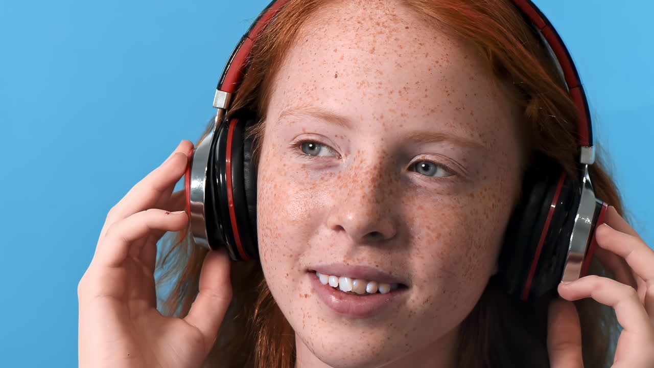 Red haired girl with lots of freckles on the face and blue eyes listening music in wireless headphones. Looking away and smile. Blue background