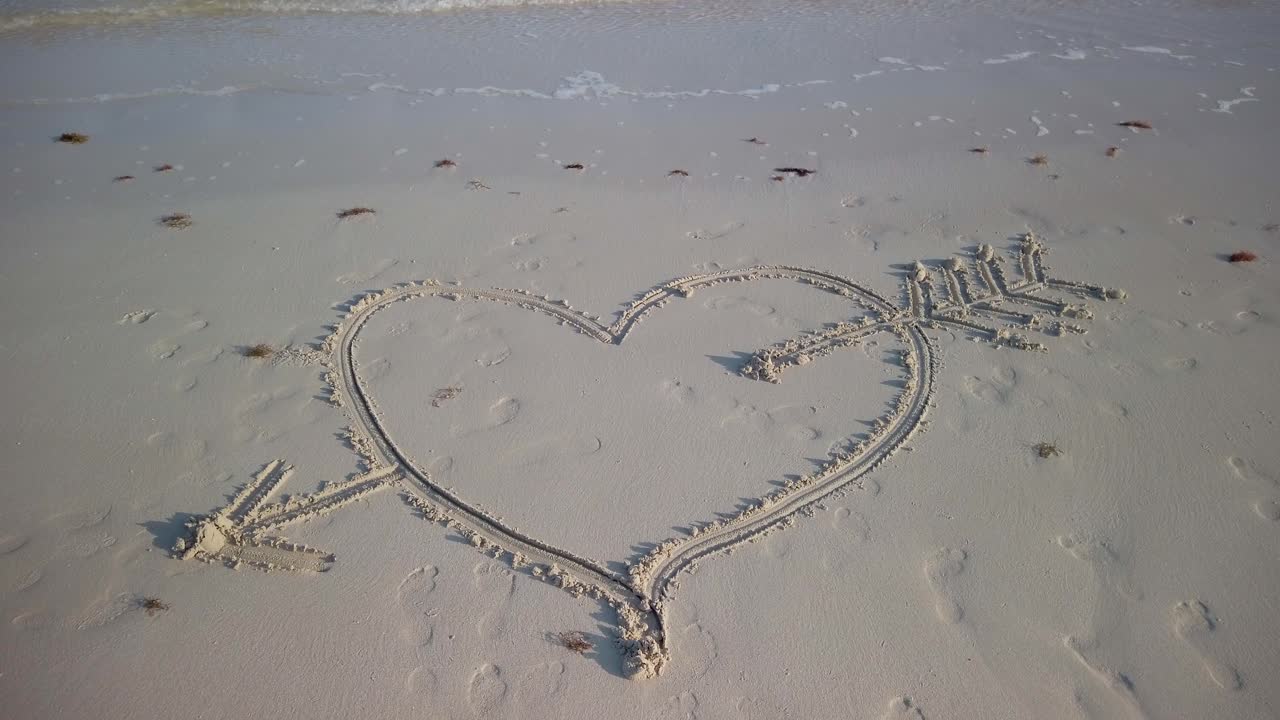 Static shot of heart with an arrow through it on a beach with water lapping on the sand