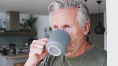 Senior-Man-Standing-And-Looking-Out-Of-Kitchen-Door-Drinking-Coffee