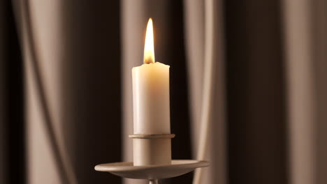 Soft-candle-burns-in-front-of-drapes-with-moving-shadows,-close-up