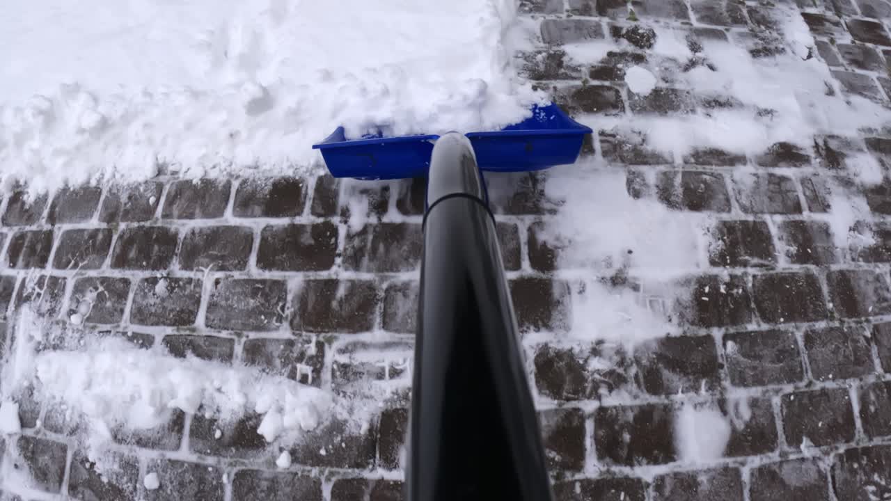 POV final pass with a blue snow shovel on cobblestone pavers, brushing off light snow dusting to reduce slip risk and refreezing on a home driveway