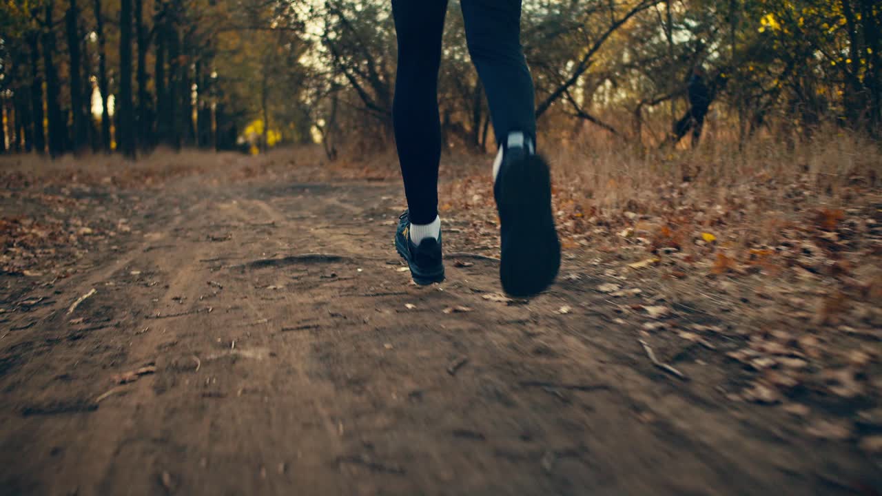 cerca de un atleta masculino en un uniforme deportivo negro y zapatillas negras corre a lo largo de un camino de tierra entre hojas caídas y hierba seca en un bosque de otoño en una mañana soleada