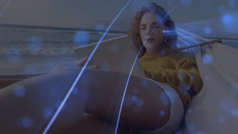 animación de manchas azules y rastros de luz sobre mujer caucásica usando tableta digital en una hamaca
