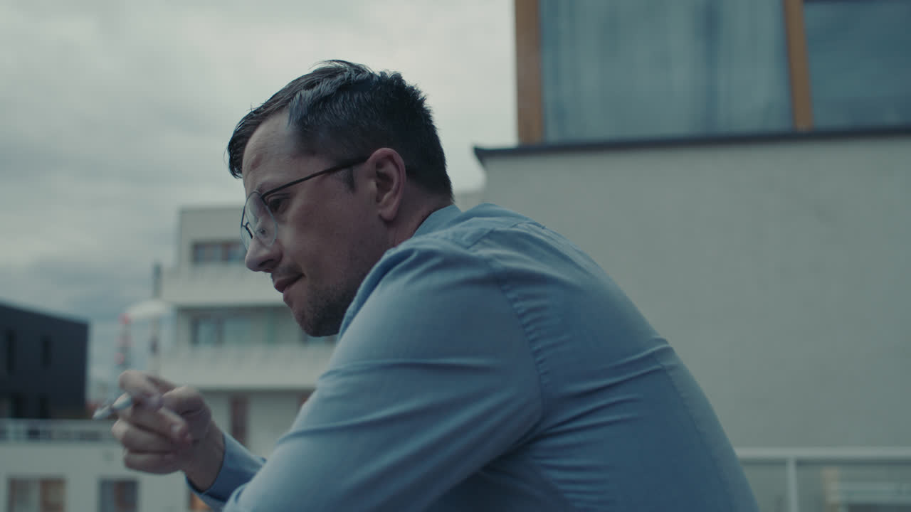 A sad man in a shirt smokes a cigarette on the balcony
