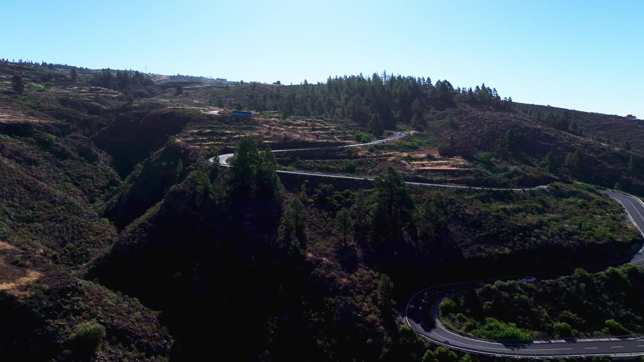 Strada di montagna tortuosa attraversa un terreno vulcanico con alberi sparsi sotto un cielo sereno.