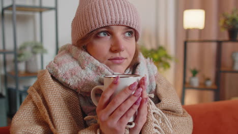 young sick woman wear hat wrapped in plaid sit alone shivering from cold on sofa drinking hot tea