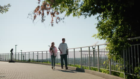 lady in pink shirt holding notebook walking alongside male friend on paved path near iron railing, engaging in cheerful conversation with greenery, cityscape, and urban architecture