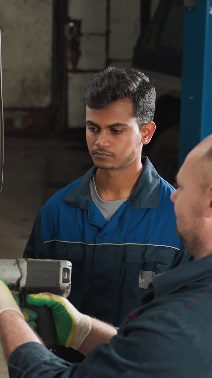 Close-up of mechanic in blue uniform using a pneumatic tool to tighten wheel bolts of a lifted car in a workshop, while a colleague attentively observes the process, ensuring precision