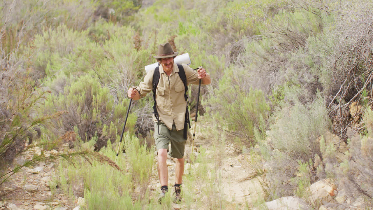 un survivalista caucásico sonriente caminando por el desierto con mochila y palos para caminar