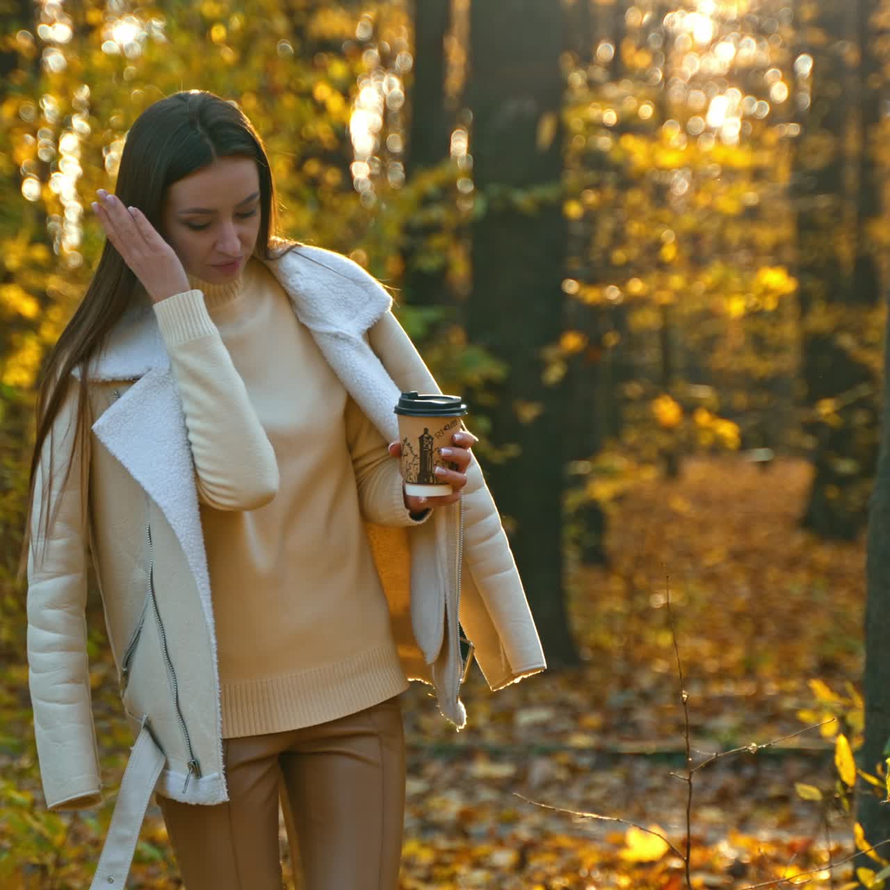 Girl walks away by the beautiful yellow carpet of leaves. Brunette turns around, arranges her hair holding a paper cup in her hand