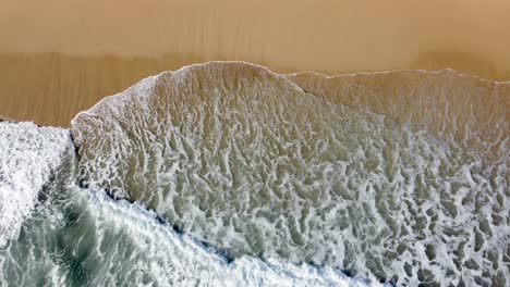 Olas-Espumosas-Rompiendo-En-Una-Orilla-Arenosa,-La-Cruda-Belleza-De-La-Naturaleza-Capturada-Desde-Arriba,-Vista-Aérea