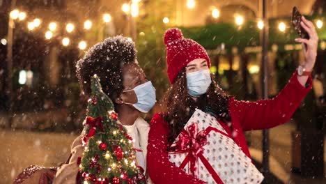 happy caucasian and african american women friends making a selfie and holding presents on the street while it¬¥s snowing in christmas