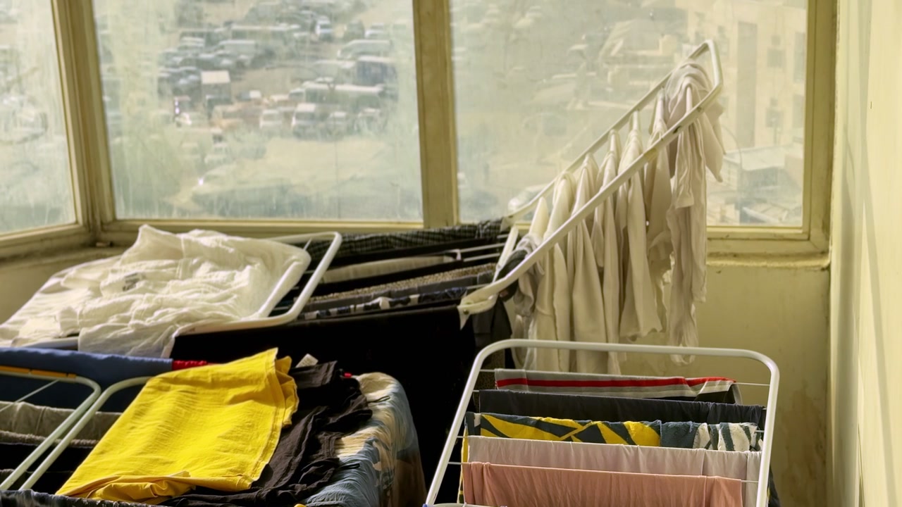 Multiple drying racks hold freshly washed clothes inside a sunlit enclosed balcony, with natural window light and a blurred urban view outside.