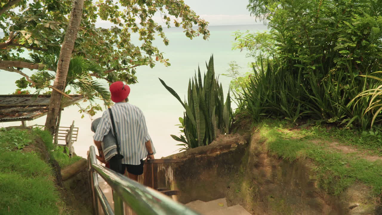 Coppia che scende gradini di sabbia verso l'oceano, uomo con cappello rosso e camicia a righe che porta un cesto di vimini mentre la donna in abito rosso lo segue, lussureggiante vegetazione tropicale e ringhiera incorniciano la riva appartata.