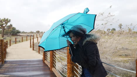 Eine-Schöne-Frau-Bei-Schlechtem-Wetter-Mit-Einem-Blauen-Regenschirm-Während-Der-Starken-Winde-Eines-Regensturms-In-Der-Zeitlupe-Der-Wüste