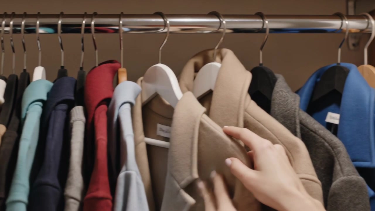 Mani che entrano nell'inquadratura, scorrendo su un appendiabiti cromato in un armadio di casa, tirando fuori un cappotto beige e ispezionando il tessuto.
