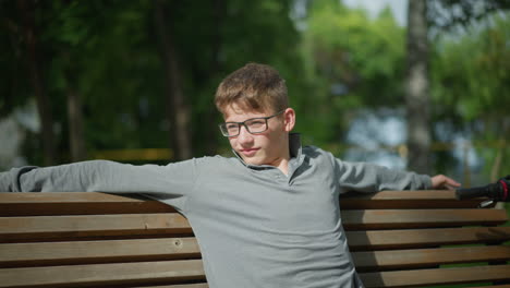 young boy wearing glasses stretches his arms wide while sitting on wooden bench, handlebar of parked bike is visible next to him, the background features trees and lush greenery