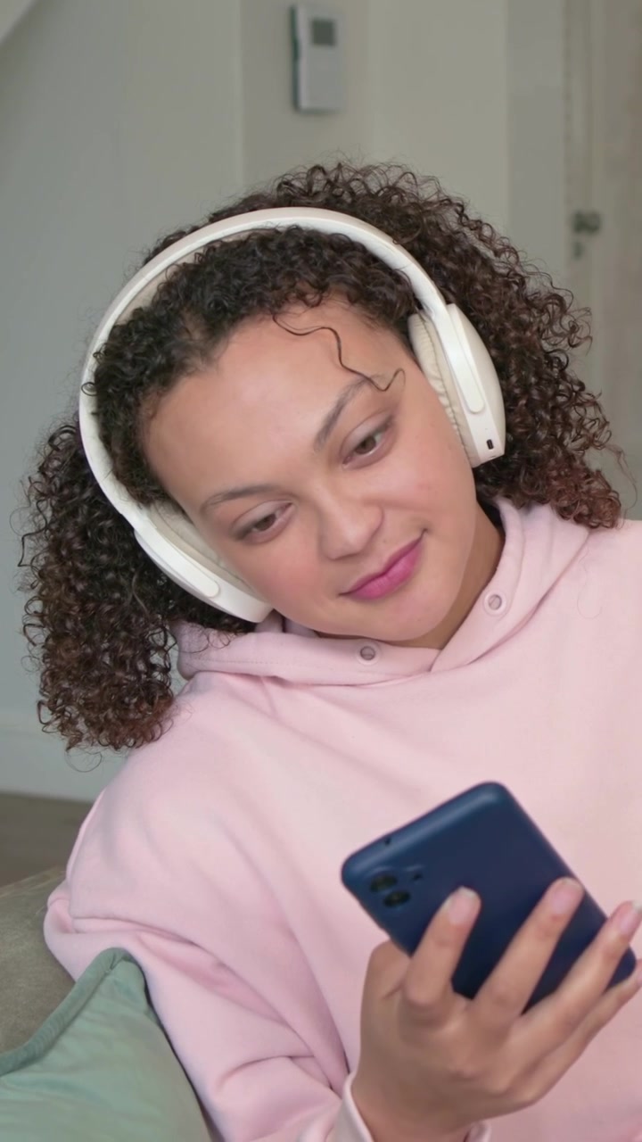 Video verticale: Una donna con felpa rosa con cappuccio scorre il telefono blu sul divano, leggendo e indossando le cuffie.