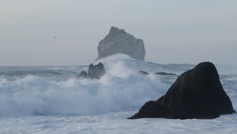 powerful white wave crashing on shallow shore with dark rock formations, slow motion